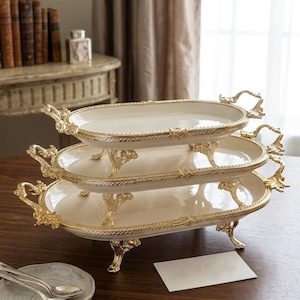 May include: A three-tiered serving tray with an off-white surface and gold-colored trim and handles. The trays are oval-shaped and rest on ornate gold-colored legs. The trays are stacked on top of each other.