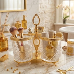 May include: A gold-toned vanity organizer with two clear, cut-glass containers. One holds makeup brushes and cotton swabs, the other a perfume bottle. The organizer has a decorative handle and sits on a marble countertop. Other gold accessories are also visible.