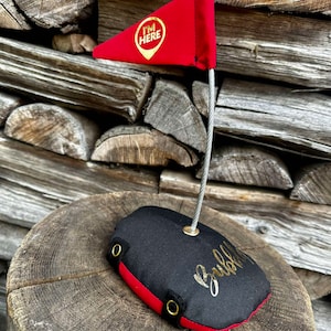 Könnte beinhalten: Eine rote dreieckige Flagge mit dem Text "I'm Here" in einem goldenen Kreis auf schwarzem Hintergrund. Die Flagge ist an einer gewichteten schwarzen und roten Basis befestigt, auf der das Wort "Bubbly" in Gold geschrieben steht.