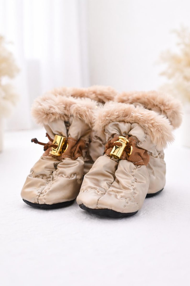 Zbliżenie na buciki dla psa Moon Dog Boots Royal w śniegu, pokazujące miękkie beżowe futerko przy cholewce oraz regulację na sznurek ze złotym stoperem. Widoczne detale wykończenia i tkaniny. Close-up of Moon Dog Boots Royal dog boots in snow, highlighting the soft beige fur trim at the cuff and the adjustable drawstring with a gold-tone stopper. The image focuses on fabric and finishing details.