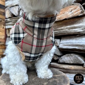 May include: A small white dog wearing a brown, black, and red plaid dog coat. The dog is sitting on a wooden surface.