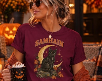 May include: A maroon short-sleeve t-shirt featuring a graphic print of a black cat sitting on a stack of books in front of a crescent moon. The text SAMHAIN is printed in a gold arched font above the cat, surrounded by autumn leaves and stars.