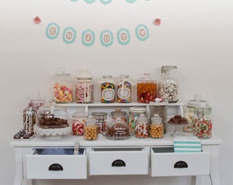 May include: A white table with three drawers and a white top. The table is decorated with jars filled with colorful candies. The jars are arranged in two rows on the table. The table is decorated with a banner that says "Candy Bar" and "Enjoy".