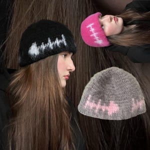 May include: Three knitted beanies in the image. One black beanie with a white zigzag design, a pink beanie with a white zigzag design, and a gray beanie with a pink zigzag design. The beanies are made of a soft, textured material.