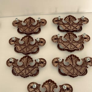 May include: Six bronze-colored drawer pulls with ornate, symmetrical designs. Each pull features a central, stylized leaf motif and a diamond-patterned base. Clear, decorative screws are attached.