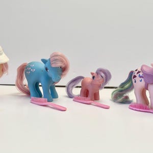 May include: Four My Little Pony figures with pink, blue, and white hair. The ponies are wearing accessories, including a hat, a saddle, and a brush. The ponies are standing on a white surface.
