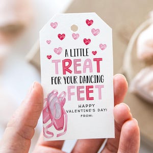 May include: A white gift tag with a Valentine's Day theme. The tag features red and pink hearts, the text "A LITTLE TREAT FOR YOUR DANCING FEET," and an illustration of pink ballet shoes. The tag also includes the text "HAPPY VALENTINE'S DAY! FROM:"