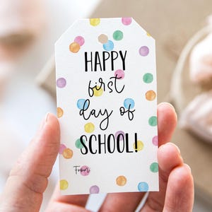 Puede incluir: Etiqueta de regalo blanca con un patrón de confeti colorido. La etiqueta dice "Happy first day of SCHOOL!" con la palabra "From" en la parte inferior.