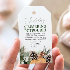 May include: A white gift tag with green text that reads "Holiday Simmering Potpourri". The tag has instructions for making potpourri and includes a watercolour illustration of pine branches, cinnamon sticks, pine cones, and orange slices.