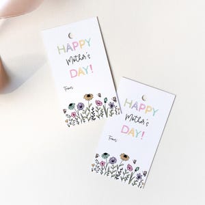 K&ouml;nnte beinhalten: Zwei wei&szlig;e Geschenkanh&auml;nger mit einem bunten Blumenrand und dem Text "Happy Mother's Day!" in Regenbogenfarben. Der Text "From:" ist unter dem Gru&szlig; gedruckt.