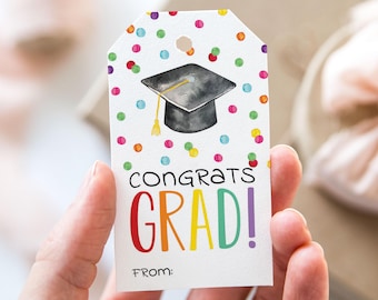 Op de afbeelding: Een afstudeergeschenktag met een aquarel illustratie van een zwarte baret en kleurrijke confetti. De tag leest "Congrats Grad!" in regenboogkleuren, met ruimte voor een "From:"-inscriptie.