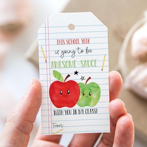 May include: A white tag with a school theme, featuring a red and green apple illustration with smiling faces. The tag reads "THIS SCHOOL YEAR is going to be AWESOME-SAUCE WITH YOU IN MY CLASS!" and includes pencil drawings.