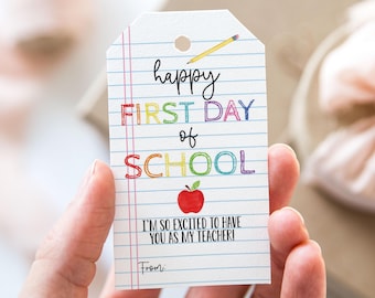 Puede incluir: Una etiqueta de regalo colorida de "Happy First Day of School" con una manzana roja y un lápiz. La etiqueta está impresa en papel blanco con un patrón de líneas azules y rojas.