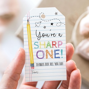 Könnte beinhalten: Ein weißer Geschenkanhänger mit einem gelben Bleistift und dem Text "You're a SHARP ONE! Thanks for all you do!" in Regenbogenfarben. Der Anhänger hat einen karierten Hintergrund und einen Platz für einen Namen unten.