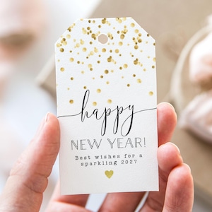 Può includere: Un'etichetta regalo bianca con coriandoli dorati e le parole "happy NEW YEAR!" in corsivo nero. L'etichetta include anche il testo "Best wishes for a sparkling 2027" e un cuore dorato.