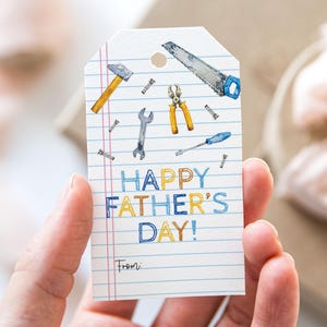 May include: A white gift tag with a watercolor illustration of tools, including a hammer, saw, and wrench. The tag reads "HAPPY FATHER'S DAY!" in blue and yellow lettering, with space to write "From:"