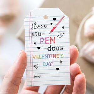 Puede incluir: Una tarjeta blanca con un dibujo de bolígrafo rosa y azul. La tarjeta dice "Have a stu-PEN-dous Valentine's Day!" con una línea para "From:"