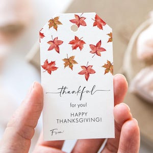 May include: A white gift tag featuring watercolour-style red and brown maple leaves. The tag reads "thankful for you! HAPPY THANKSGIVING!" with space for "From:" at the bottom. The tag is held in a person's hand.