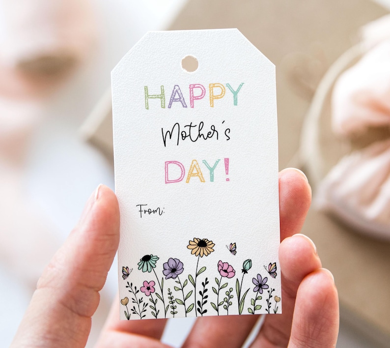 K&ouml;nnte beinhalten: Ein wei&szlig;er Geschenkanh&auml;nger mit einer bunten "Happy Mother's Day!"-Botschaft und einem floralen Rand. Der Anh&auml;nger hat Platz f&uuml;r eine handgeschriebene Nachricht und steht unten "Von:"