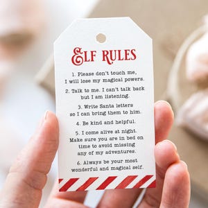 May include: A white gift tag with the text "ELF RULES" in red script. The tag lists six rules in black text, with a red and white diagonal stripe design at the bottom. The tag is held by a hand.