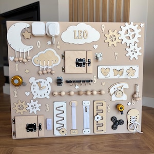 Op de afbeelding: Een beige houten activiteitenbord voor peuters, met diverse interactieve elementen. Inclusief tandwielen, grendels, een klok, een telefoon en de naam "LEO" in witte letters. Ontworpen om de fijne motoriek te stimuleren.