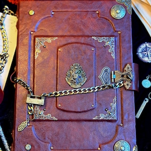 Puede incluir: Un diario rectangular encuadernado en cuero rojo oscuro con protectores de esquina de latón de estilo antiguo, tachuelas decorativas circulares y un emblema central del escudo de Hogwarts. El libro se asegura con un cierre.