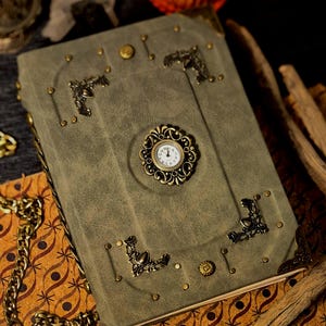 Op de afbeelding: Een vintage-stijl notitieboek met een saliegroene kaft, versierd met sierlijke messing hoekaccenten, studs en een decoratieve klok. Een gouden ketting is over de zijkant gedrapeerd. Het notitieboek heeft een klassieke, antieke uitstraling.
