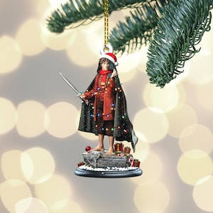 May include: A Christmas ornament depicting a character from The Lord of the Rings, wearing a Santa hat and holding a sword. The figure is dressed in a red and black outfit, with a black cape and a string of lights. The ornament is hanging from a gold string.