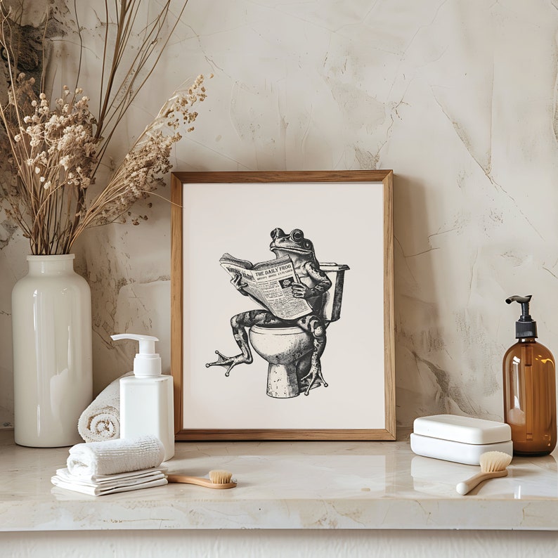 K&ouml;nnte beinhalten: Eine Schwarzwei&szlig;illustration eines Frosches, der auf einer Toilette sitzt und eine Zeitung mit dem Titel "The Daily Frog" liest. Die Illustration ist in einem braunen Holzrahmen gerahmt und an einer wei&szlig;en Wand aufgeh&auml;ngt.