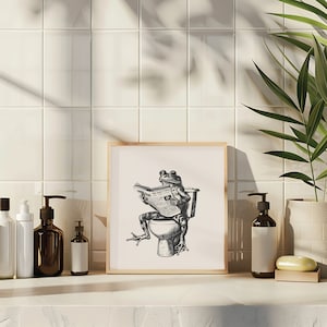 K&ouml;nnte beinhalten: Eine Schwarzwei&szlig;illustration eines Frosches, der auf einer Toilette sitzt und eine Zeitung mit dem Titel "The Daily Frog" liest. Die Illustration ist in einem hellen Holzrahmen gerahmt und h&auml;ngt an einer wei&szlig;en gekachelten Wand.