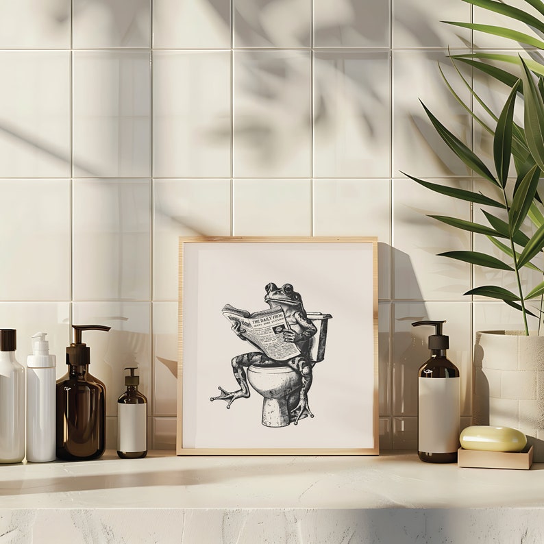 K&ouml;nnte beinhalten: Eine Schwarzwei&szlig;illustration eines Frosches, der auf einer Toilette sitzt und eine Zeitung mit dem Titel "The Daily Frog" liest. Die Illustration ist in einem hellen Holzrahmen gerahmt und h&auml;ngt an einer wei&szlig;en gekachelten Wand.