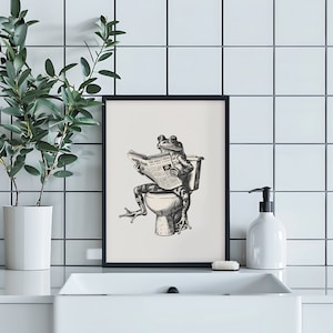 K&ouml;nnte beinhalten: Schwarzwei&szlig;-Illustration eines Frosches, der auf einer Toilette sitzt und eine Zeitung mit dem Titel "The Daily Frog" liest. Die Illustration ist in einem schwarzen Rahmen gerahmt und an einer wei&szlig;en gekachelten Wand aufgeh&auml;ngt.