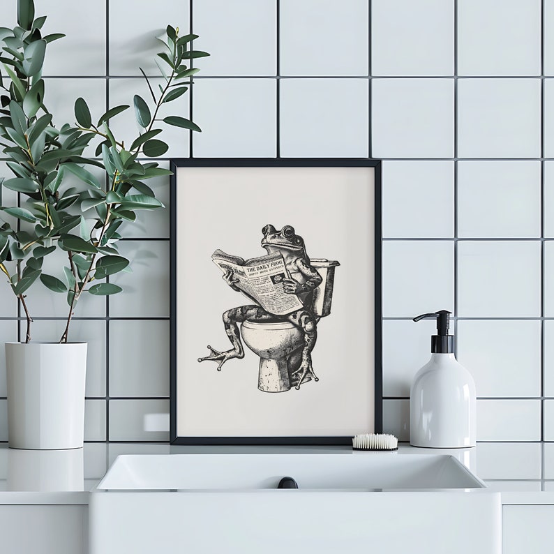 K&ouml;nnte beinhalten: Schwarzwei&szlig;-Illustration eines Frosches, der auf einer Toilette sitzt und eine Zeitung mit dem Titel "The Daily Frog" liest. Die Illustration ist in einem schwarzen Rahmen gerahmt und an einer wei&szlig;en gekachelten Wand aufgeh&auml;ngt.