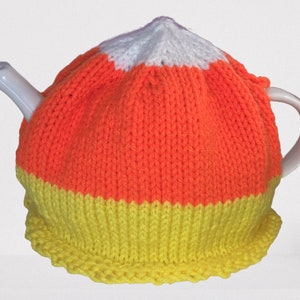 May include: A white ceramic teapot covered with a knitted tea cozy. The cozy is designed to resemble a candy corn, with sections of white, orange, and yellow. The cozy fits snugly over the teapot, with a scalloped edge at the bottom.