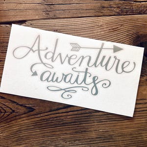 May include: A white rectangular sticker with the words "Adventure awaits" in a cursive font, with an arrow graphic. The sticker is light gray and sits on a wooden surface.