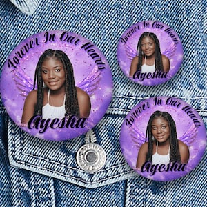 Puede incluir: Tres botones redondos morados con el texto "Forever In Our Hearts" y el nombre "Ayesha". Cada botón presenta una foto de una mujer con alas de ángel. Los botones están sobre un fondo de mezclilla.