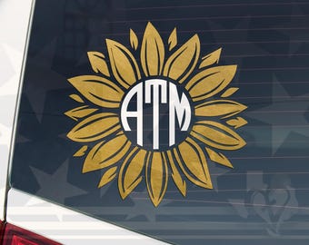 Könnte beinhalten: Ein goldfarbener Sonnenblumenaufkleber mit einem weißen Monogramm der Buchstaben "ATM" in der Mitte. Der Aufkleber wird auf einer Autoscheibe mit dunklem Hintergrund angezeigt.