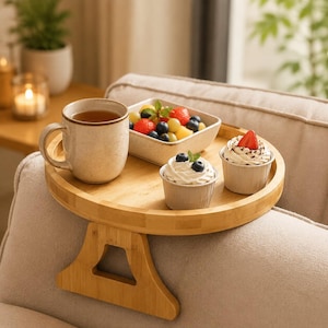 Puede incluir: Una bandeja redonda de bambú para el brazo del sofá con base de pinza. Sobre la bandeja hay una taza de cerámica beige moteada con té, un cuenco cuadrado blanco con frutos rojos y dos vasos de papel con nata montada y fruta.