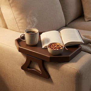 Puede incluir: Una bandeja hexagonal marrón para el brazo del sofá sostiene una taza de café humeante, un libro abierto y un pequeño cuenco con aperitivos. La bandeja está sostenida por un soporte que se adapta al brazo del sofá. La escena sugiere un rincón de lectura acogedor.