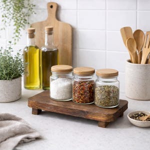 May include: A kitchen scene featuring glass jars with wooden lids filled with spices, displayed on a wooden tray. Olive oil bottles and wooden utensils are in the background. A small potted plant and a cutting board are also visible.
