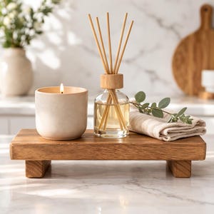 May include: A burning candle in a beige ceramic pot, a glass diffuser with wooden reeds, and a folded beige and white striped cloth are displayed on a small, rectangular wooden tray. A sprig of greenery adds a natural touch.