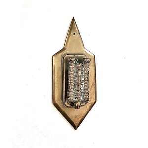 May include: A vintage brass wall hanging with a detailed castle relief. The castle is set within a rectangular frame, mounted on a brass shield shape with a pointed top. It has a small hole at the top for hanging, typical of decorative items.