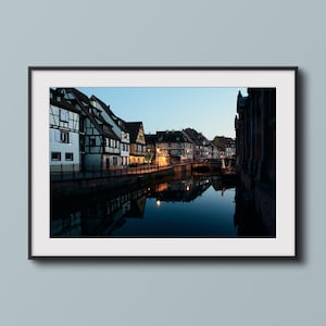 Può includere: Vista di un canale in una città europea al crepuscolo. Il canale è fiancheggiato da edifici tradizionali con facciate a graticcio. Gli edifici si riflettono nell'acqua, creando un'immagine simmetrica.