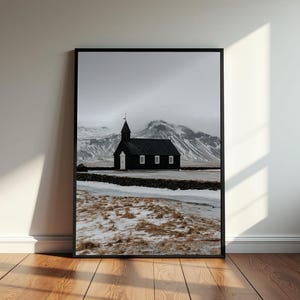 Può includere: Una chiesa nera con una porta bianca si erge in un campo innevato con una catena montuosa sullo sfondo.