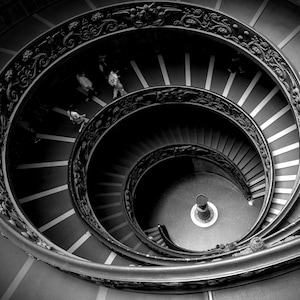 以下が含まれることがあります： 螺旋階段を人が上っている白黒写真。階段は石造りで、手すりは鉄製です。
