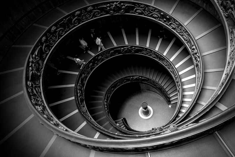 以下が含まれることがあります： 螺旋階段を人が上っている白黒写真。階段は石造りで、手すりは鉄製です。