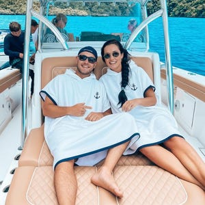 May include: Two white hooded poncho towels with dark blue trim and a small black anchor graphic on the chest. The ponchos are made of absorbent fabric and feature a loose, comfortable fit with short sleeves, designed for drying off after swimming.