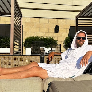 May include: A white hooded towel poncho featuring a loose-fitting design with short sleeves and a front pocket. The garment is made of a soft, absorbent fabric and provides full coverage, extending to the mid-thigh area for comfort and utility.