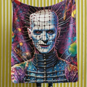 Könnte beinhalten: Eine bunte Kuscheldecke mit einer grafischen Illustration eines Horrorfilmcharakters mit einem Gitter aus Metallhaken und einem bedrohlichen Ausdruck.