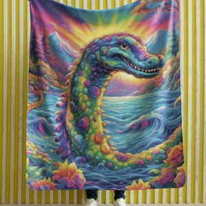 May include: A colorful fleece blanket featuring a psychedelic sea monster with a bright green, blue, and yellow pattern. The monster is emerging from a body of water with a sunset in the background.